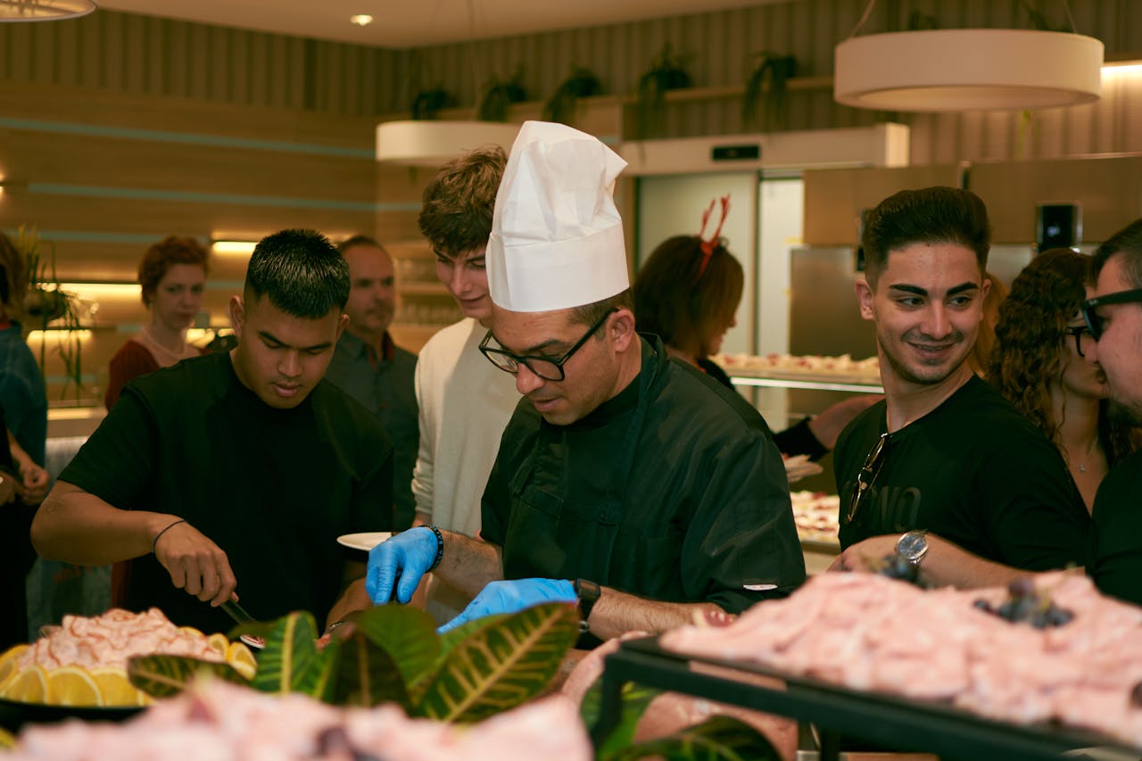 services-02 A chef in glasses serves food to guests at a lively indoor buffet event.